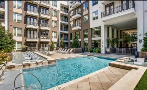 Resort-style swimming pool with spa, surrounded by lounge chairs and modern apartment buildings in an urban courtyard setting.
