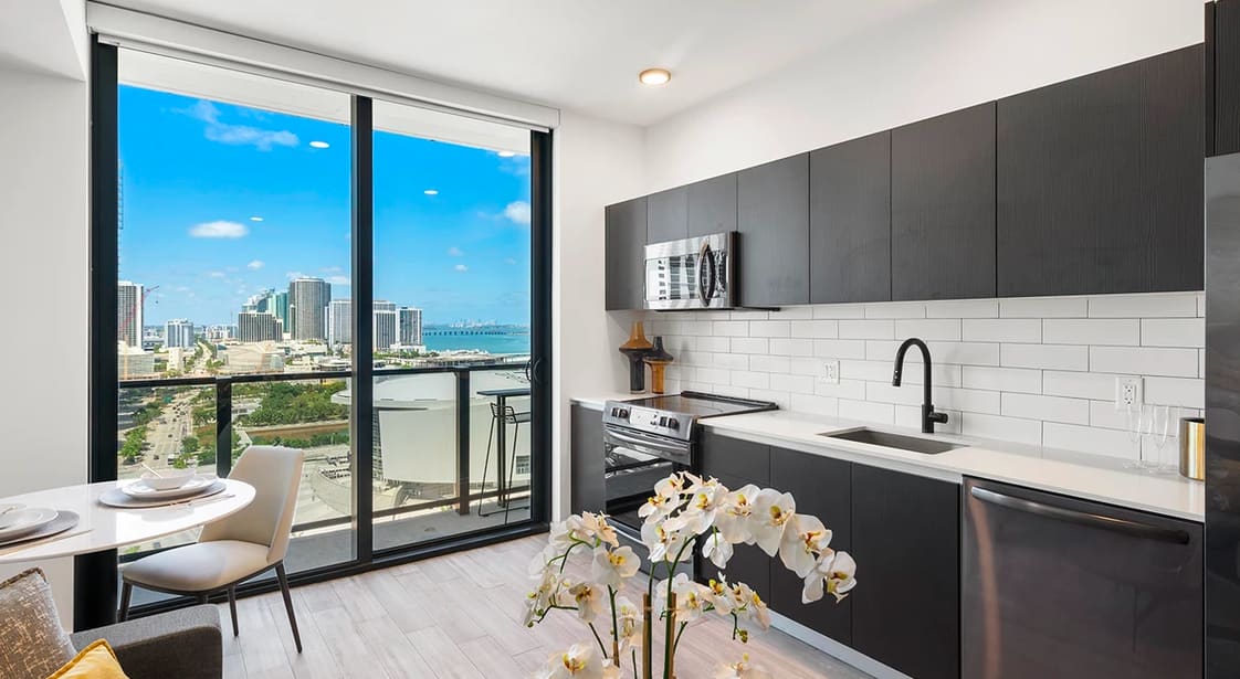 Modern kitchen with black cabinetry, white subway tile backsplash, and floor-to-ceiling windows showing Miami skyline and ocean views.