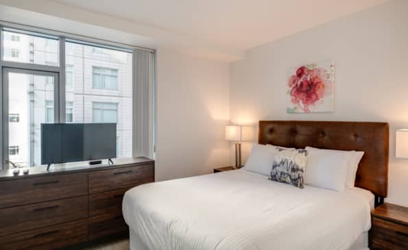 Bedroom with queen bed, brown wood headboard and dresser, flat-screen TV, large window overlooking city buildings, and bedside lamps.