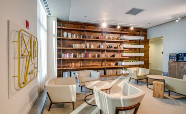 Contemporary lounge with cream armchairs, wooden shelving wall, yellow geometric wall art, and large windows with natural light.
