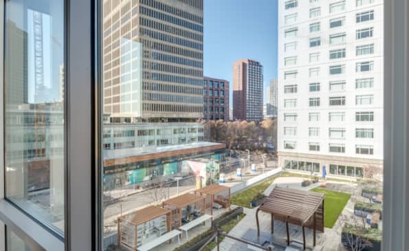 High-rise window view of Cambridge cityscape with downtown buildings, rooftop terrace with seating below, and clear blue sky.