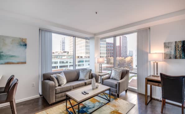 Corner living room with gray sofa, armchair, coffee table, and expansive windows showing downtown Cambridge buildings and sky.