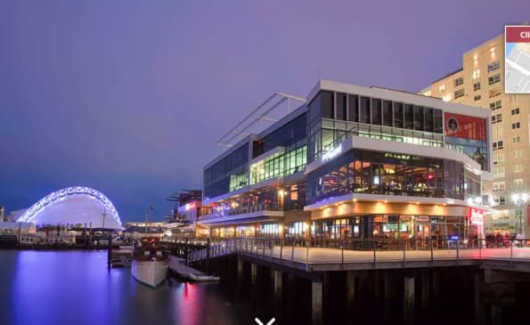 Modern waterfront building at dusk with illuminated storefronts, adjacent to a lit Ferris wheel and calm harbor water.
