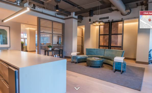 Open-concept apartment interior with white kitchen island, teal curved sofa, glass partition walls, and polished concrete flooring.