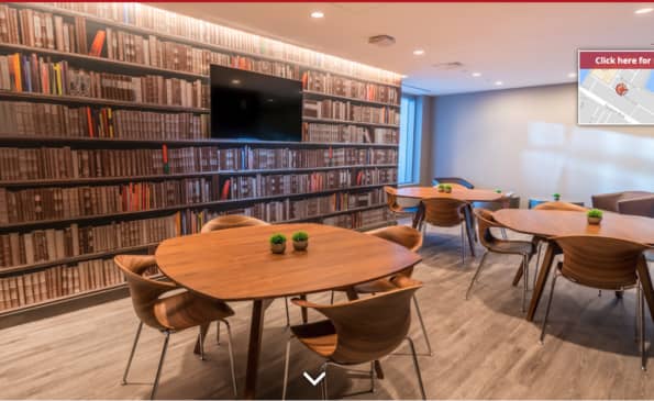 Shared lounge with wall-to-wall bookshelves, wooden dining tables, mid-century chairs, and recessed lighting.