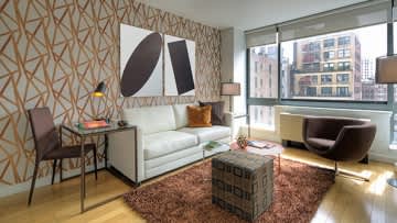 Studio apartment with cream sofa, geometric patterned accent wall, small desk, and floor-to-ceiling windows overlooking city buildings.