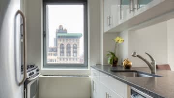 Small kitchen with white cabinetry, gray countertop, and large window overlooking historic New York City building.