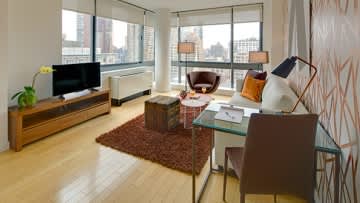 Modern living room with wooden TV console, brown sofa, glass coffee table, and large windows overlooking city skyline.