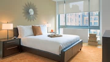 Bedroom with queen bed, dark wood frame, white bedding, sunburst wall clock, two nightstands with lamps, and large windows overlooking the city.