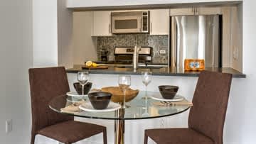 Compact kitchen with stainless steel refrigerator and microwave, round glass dining table with two brown chairs, white countertop with bar seating.