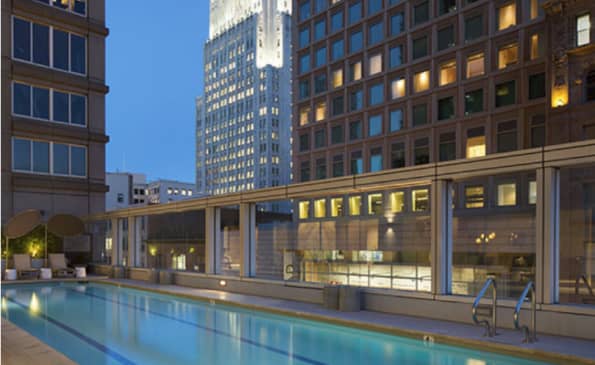 Rooftop swimming pool at dusk with illuminated downtown San Francisco skyscrapers and glass-walled building behind it.