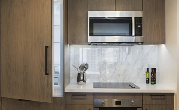 Compact kitchen with marble backsplash, stainless steel microwave, white countertop, and wood cabinetry with black handles.
