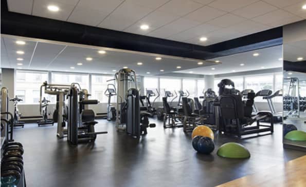 Modern gym with cardio machines, weight equipment, medicine balls, and recessed ceiling lights in a spacious room with large windows.