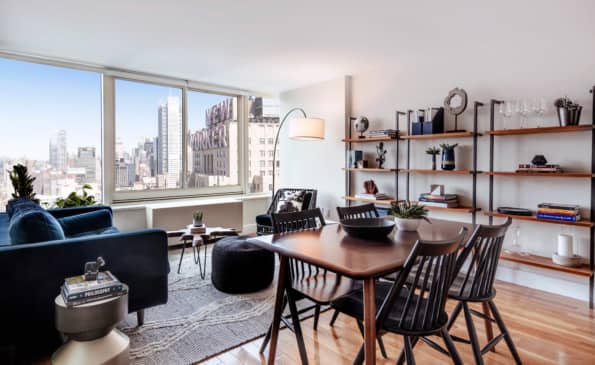 Modern apartment living room with dining table, black chairs, wooden shelving, arc lamp, and expansive windows showing Manhattan skyline views.