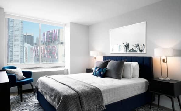 Modern bedroom with navy bed, white bedding, two nightstands with lamps, and large windows showing New York City skyline and street-level signage.