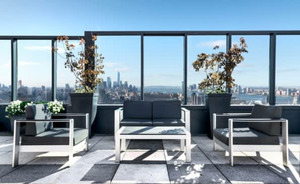 Rooftop terrace with white and black modern furniture, potted plants, and expansive floor-to-ceiling windows overlooking the Manhattan skyline.