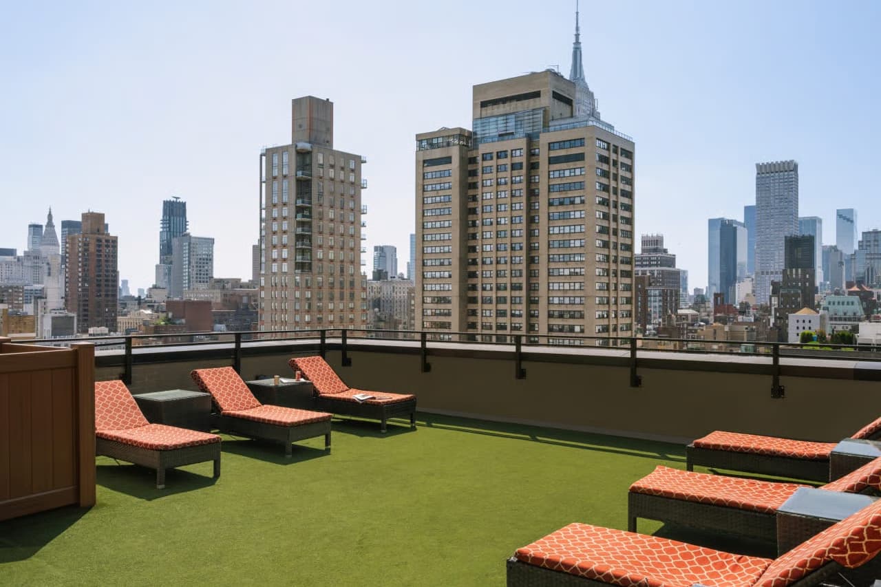 Rooftop terrace with orange cushioned lounge chairs on artificial turf, metal railing, and Manhattan skyline in background.