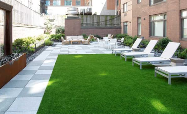 Rooftop terrace with artificial grass, white lounge chairs on right, paved seating area with tables and chairs in background, surrounded by brick buildings.