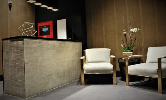Modern lobby with woven reception desk, two cream upholstered armchairs with wooden frames, and decorative objects on dark shelving.