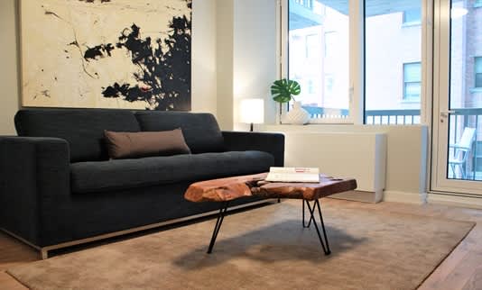 Living room with black sofa, wooden coffee table, large abstract artwork, and floor-to-ceiling windows with city views.