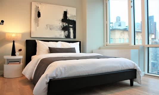 Bedroom with black bed frame, white and gray bedding, abstract wall art, nightstand with lamp, and large windows with city views.