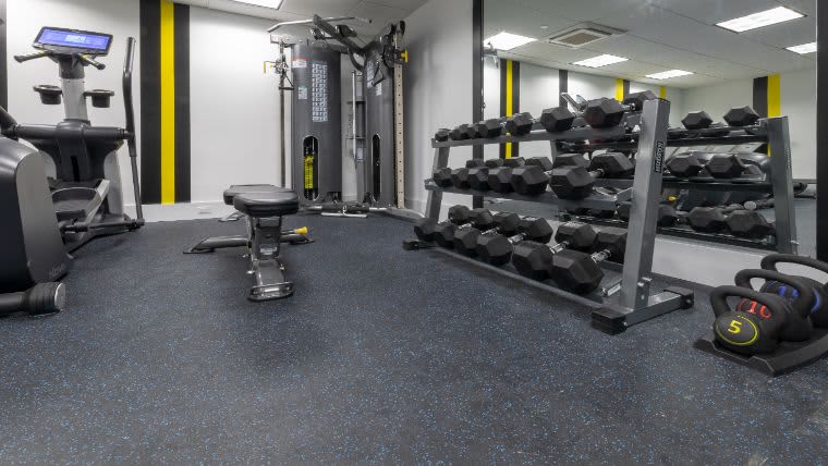Modern gym with cardio machine on left, dumbbell racks along right wall, weight bench in center, and blue speckled flooring.