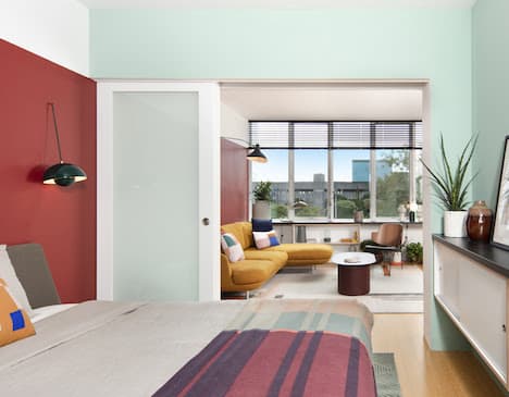 Bedroom with striped area rug opening to living room with yellow sofa, large windows, and city views.