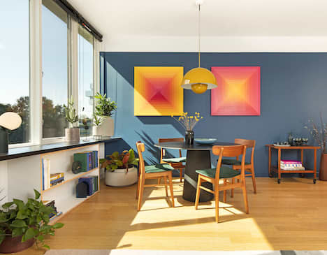 Dining room with wooden table and chairs, blue accent wall with colorful abstract art, pendant light, and large windows overlooking greenery.