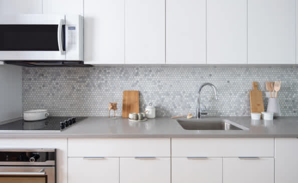 White kitchen with gray quartz countertop, stainless steel sink, geometric tile backsplash, and white cabinetry with microwave and stovetop.