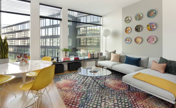 Open-concept living room with white sofa, yellow accent chair, geometric patterned rug, and floor-to-ceiling windows overlooking the city.