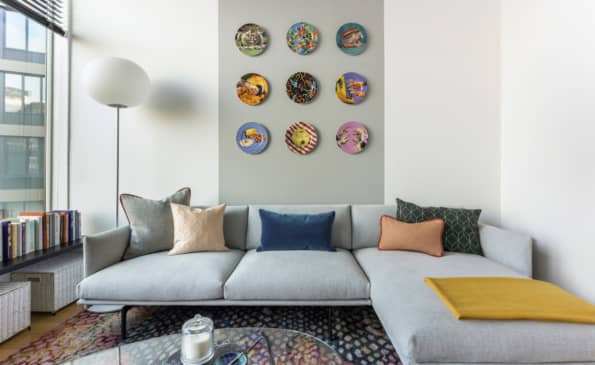 Living room with gray sectional sofa, colorful throw pillows, nine decorative plates on wall, floor lamp, and city views through windows.