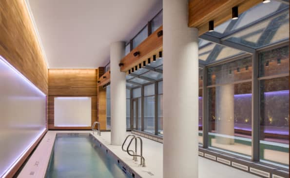 Indoor swimming pool with ambient purple lighting, large windows overlooking the city, white columns, and wooden ceiling details.
