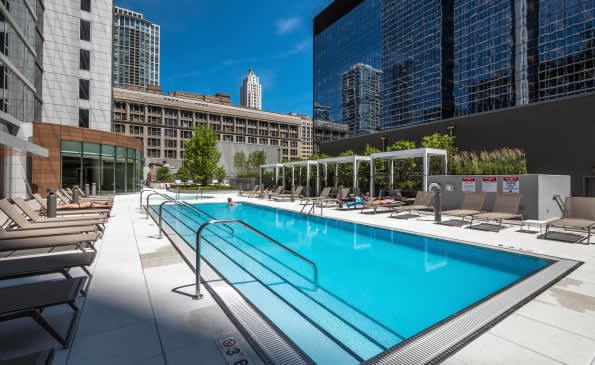 Rooftop swimming pool with lane markers, lounge chairs, and Chicago skyline in background under clear blue sky.