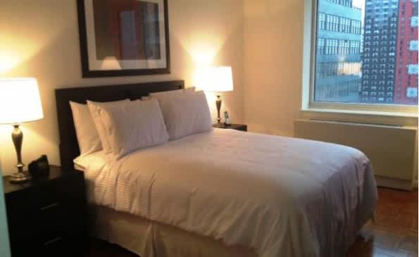 Queen bedroom with white bedding, black headboard, two table lamps, and window overlooking downtown Manhattan buildings.