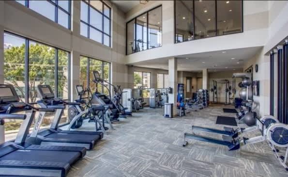 Modern fitness center with treadmills, exercise bikes, and weight equipment arranged on patterned flooring beneath high ceilings and large windows.