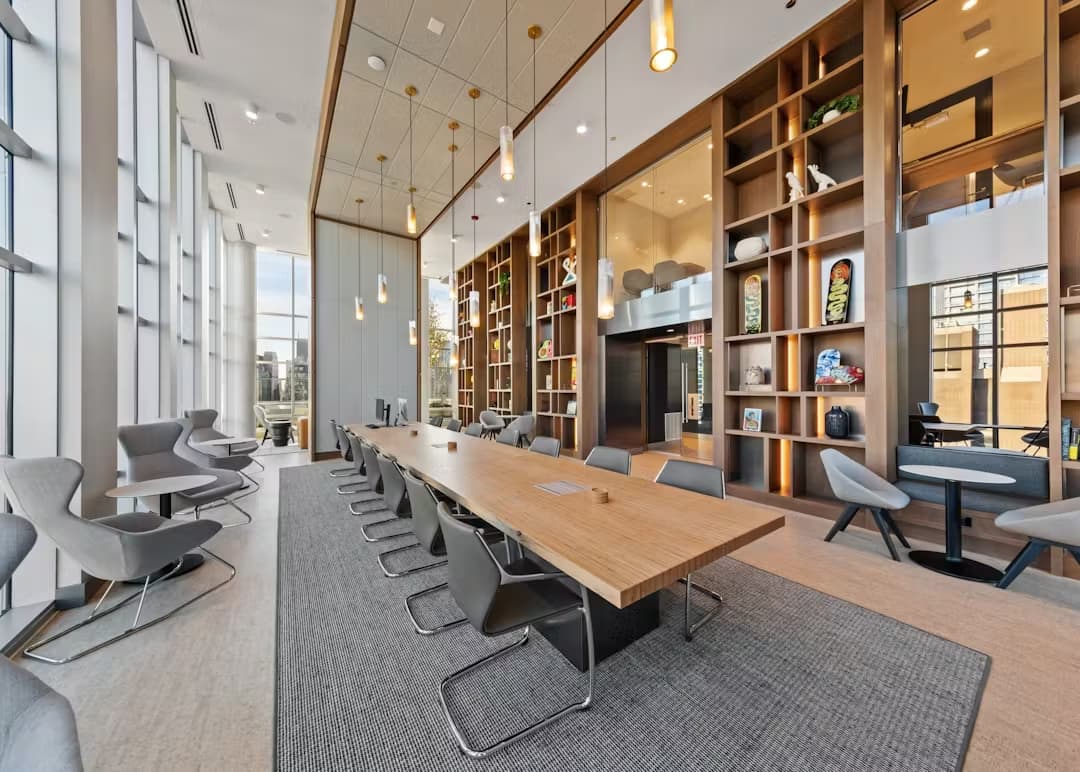 Open-plan common room with long wood dining table, gray chairs, floor-to-ceiling wood shelving, pendant lights, and large windows.