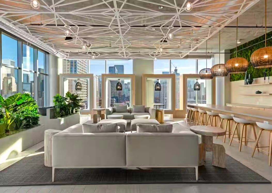 Modern lounge with gray seating, geometric ceiling, hanging copper pendants, floor-to-ceiling windows with city views, and bar seating.