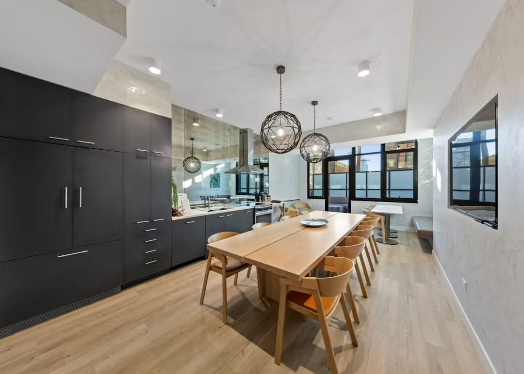 Open-concept kitchen and dining room with black cabinetry, light wood dining table, six wooden chairs, geometric pendant lights, and floor-to-ceiling windows.