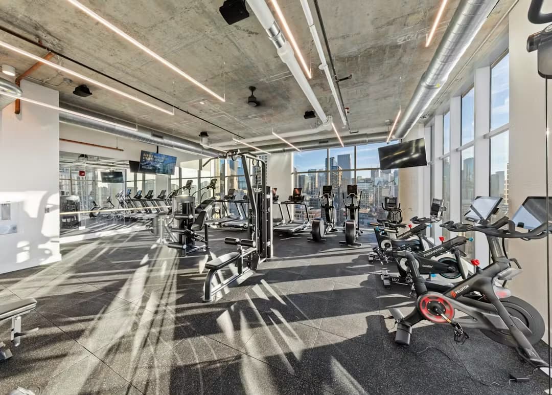 Modern gym with cardio bikes, treadmills, and strength machines; floor-to-ceiling windows with Chicago skyline views; concrete ceiling with linear lighting.
