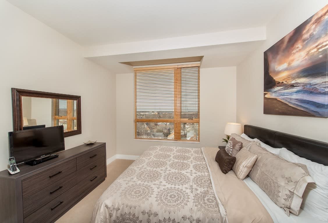 Bedroom with queen bed, dark wood dresser and TV stand, large windows with wooden blinds, and framed ocean artwork on wall.