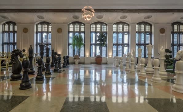 Spacious lobby with oversized black and white chess pieces, tall arched windows, polished floor, and classical architectural elements.