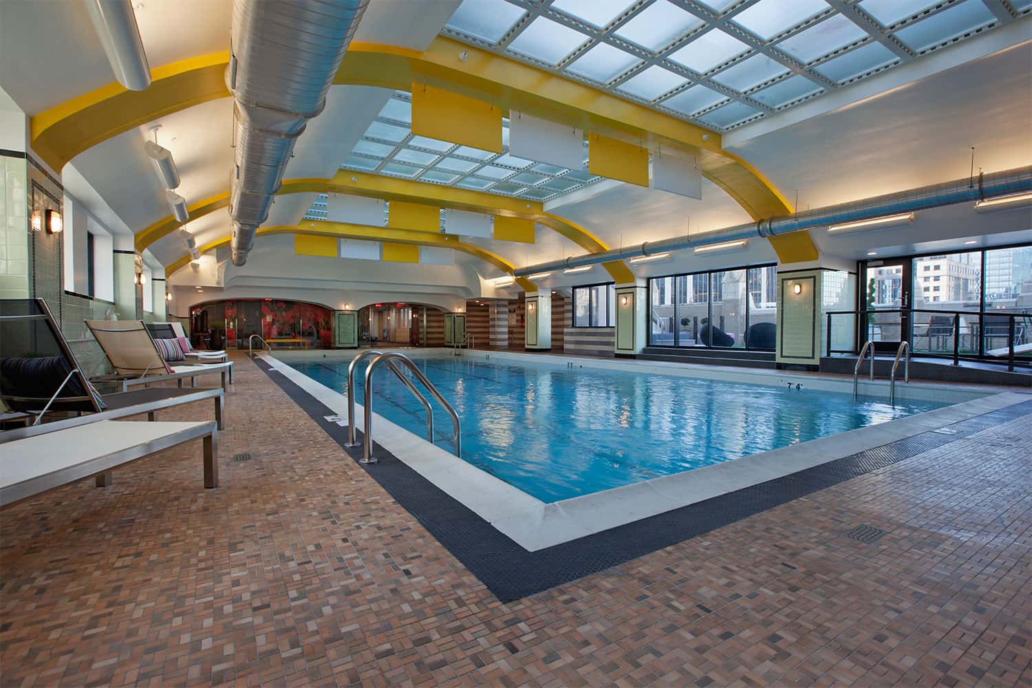 Indoor swimming pool with blue water, white deck edge, metal handrails, lounge chairs, yellow ceiling beams, skylights, and Chicago skyline visible through windows.