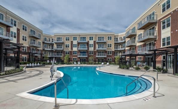 Curved resort-style swimming pool with lounge chairs, surrounded by multi-story brick and tan apartment buildings in a courtyard setting.