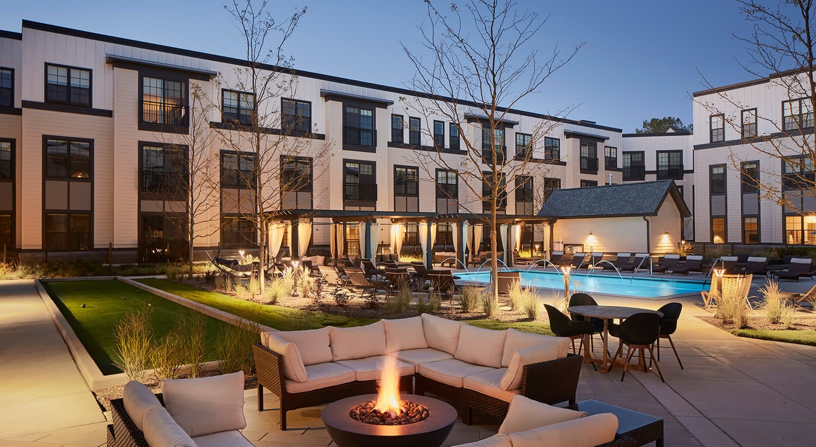 Courtyard fire pit with beige cushioned seating, lit pool, and multi-story residential buildings at dusk.