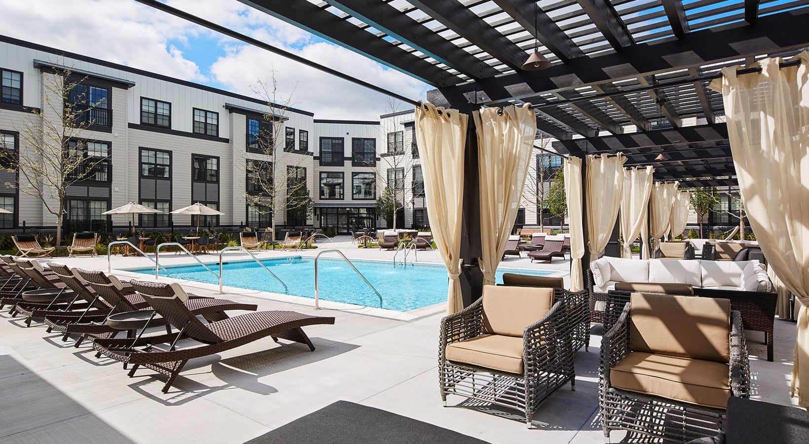 Resort-style pool with lounge chairs, cream pergola with draped curtains, wicker seating areas, and multi-story residential buildings surrounding the courtyard.