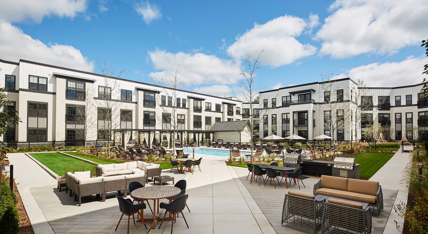 Courtyard with pool, lounge seating, dining tables, and modern white apartment buildings under blue sky.