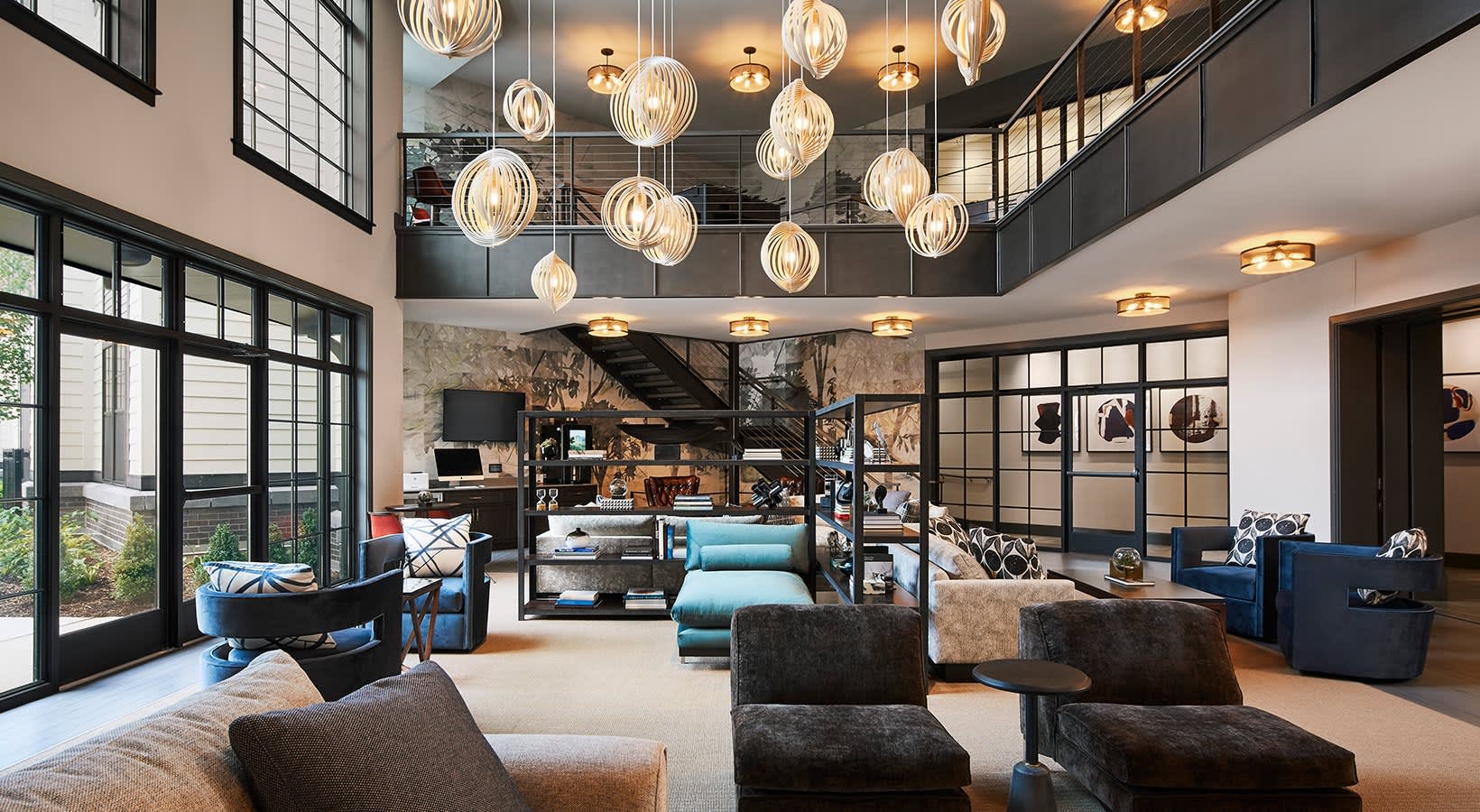 Two-story loft living room with sculptural pendant lights, dark upholstered seating, glass railings, and floor-to-ceiling windows overlooking outdoor space.