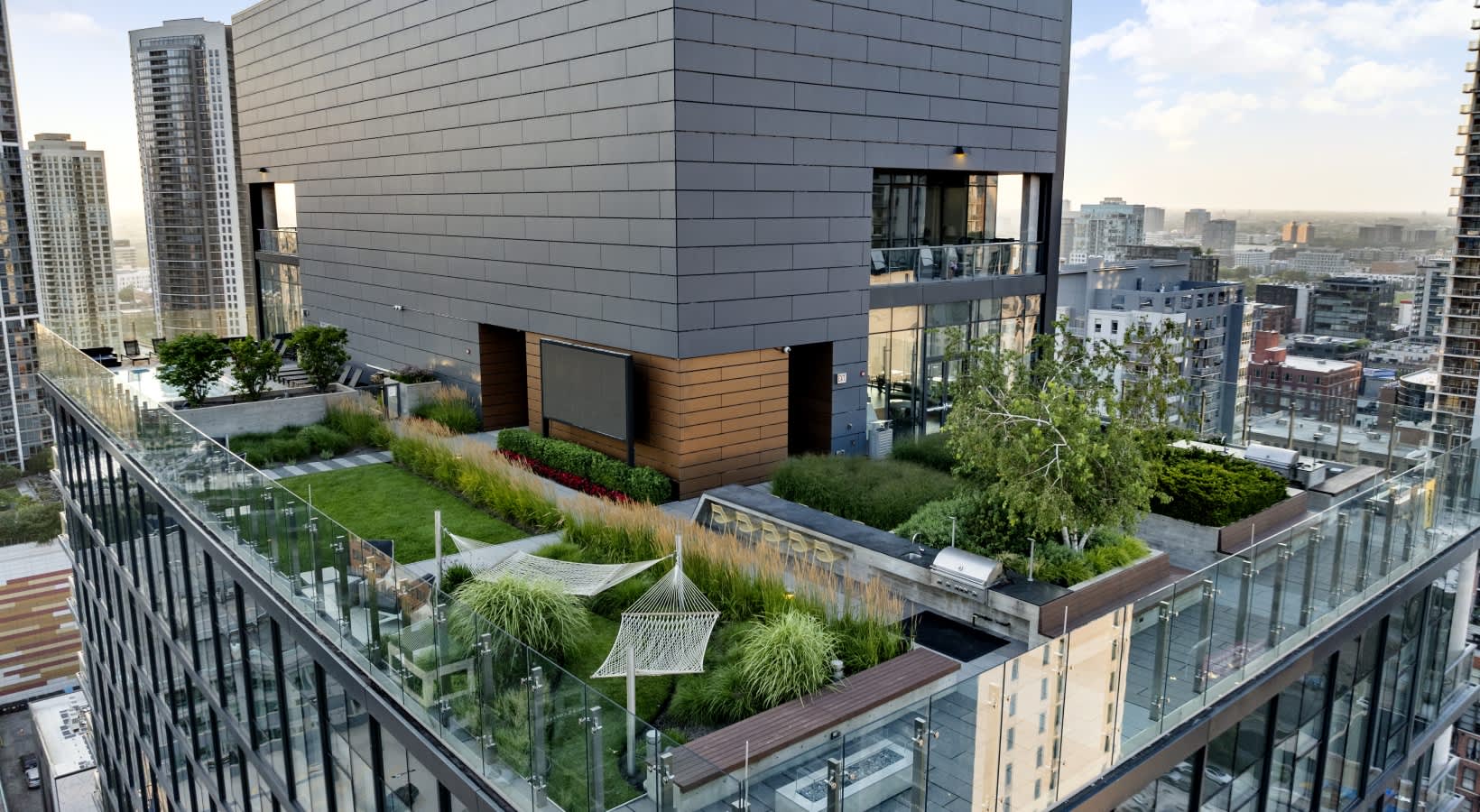 Rooftop terrace with ornamental grasses, green lawn, hammock, and glass railings overlooking Chicago skyline and surrounding buildings.