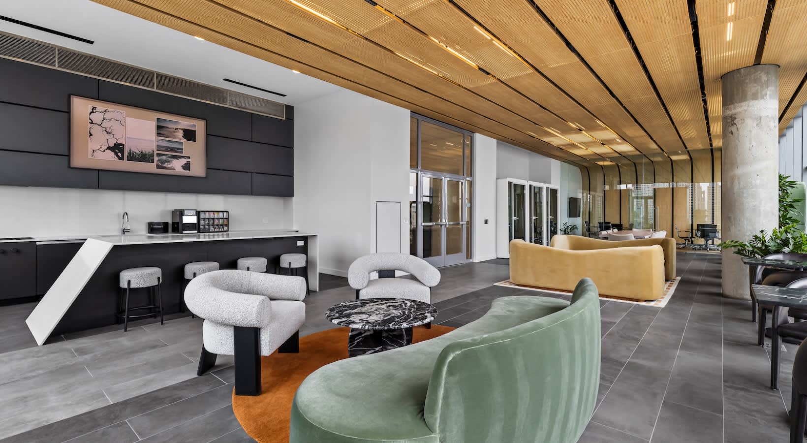 Open-plan lounge with curved white and green upholstered chairs, yellow sofa, black kitchen counter, and wood-paneled ceiling with skylights.