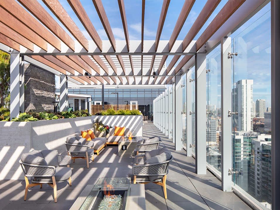 High-rise rooftop terrace with wood pergola, modern lounge chairs with yellow cushions, fire pit, planters, and floor-to-ceiling glass walls overlooking downtown Chicago.